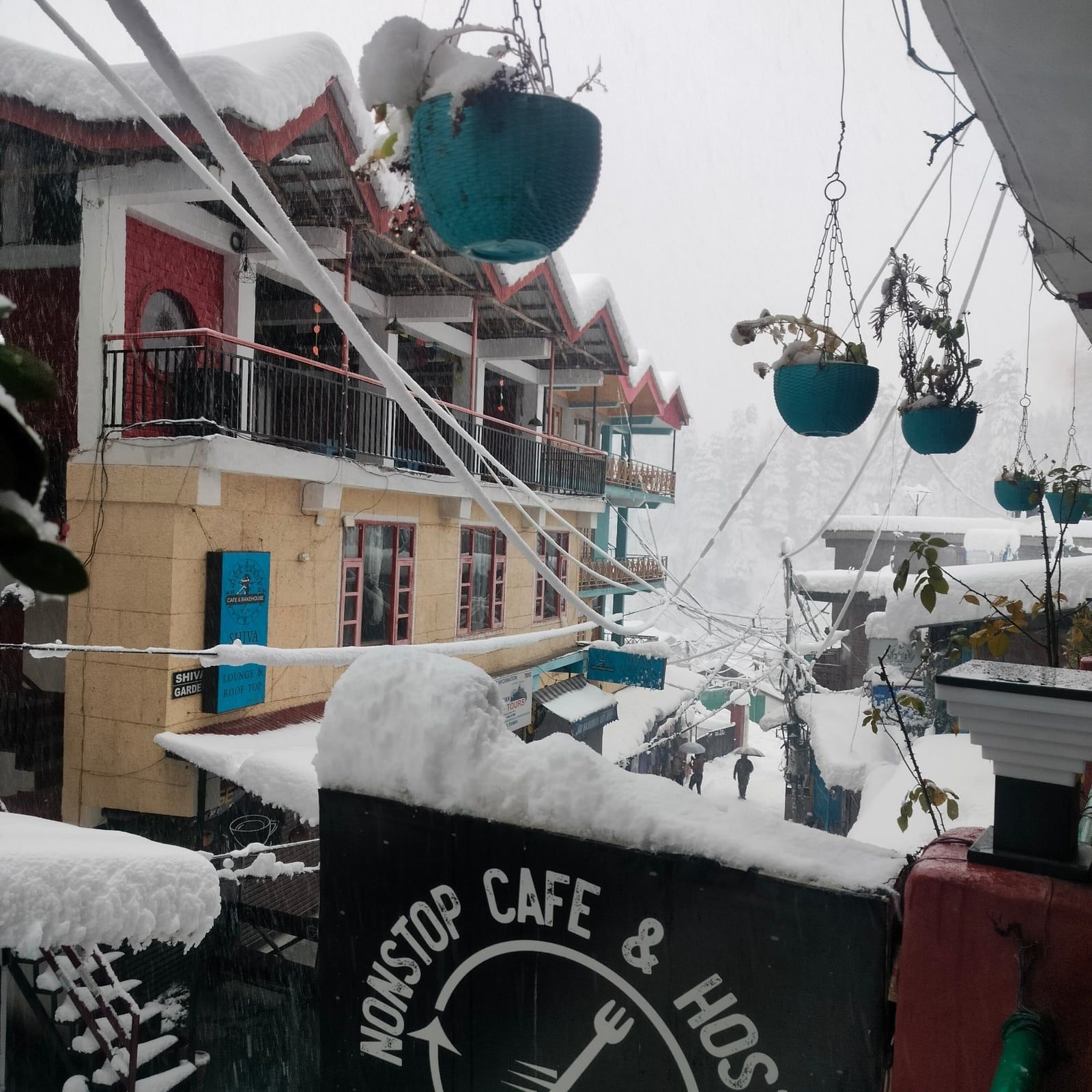 Snow on the Non Stop Café & Hostel sign and the rooftops beyond, with hanging plant pots
